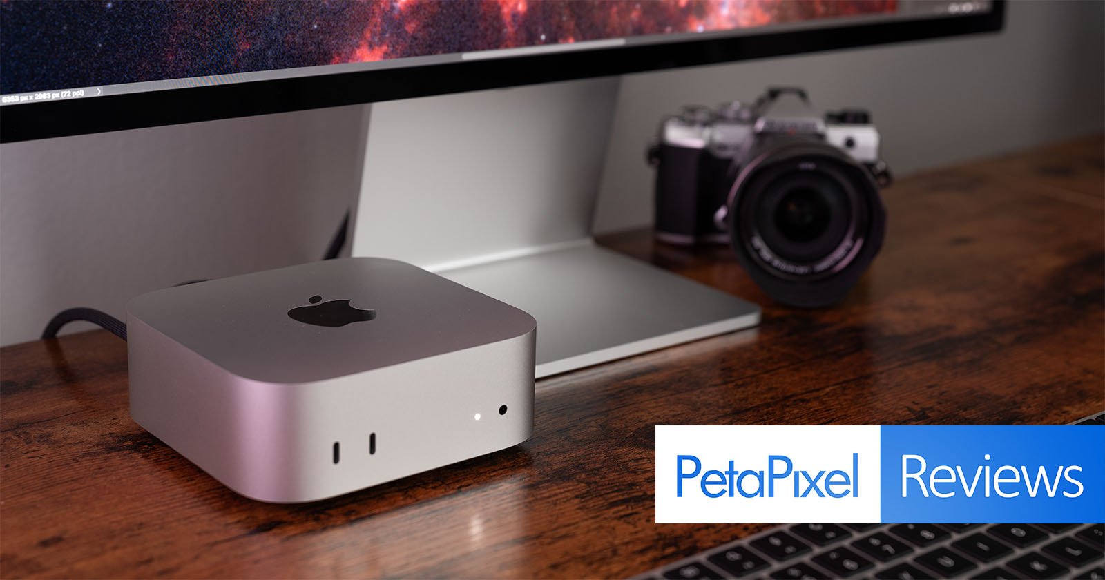A sleek silver computer device with an apple logo is placed on a wooden desk in front of a large monitor. A digital camera and a keyboard are nearby. The text PetaPixel Reviews is overlaid on the image.