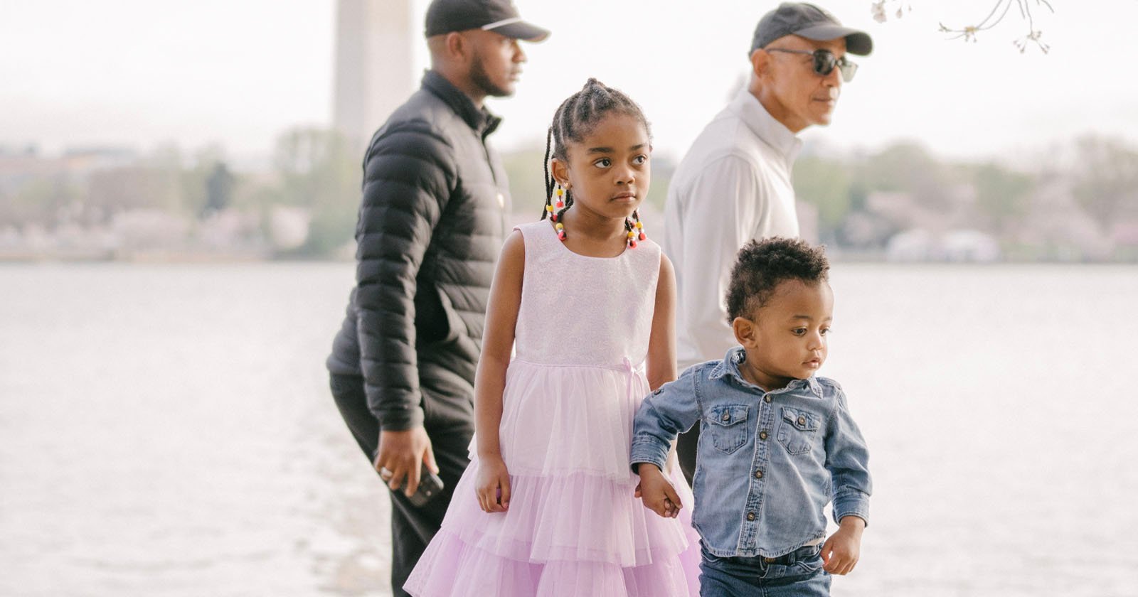 Two children, a girl in a white and pink dress and a boy in a denim outfit, stand by water. Two adults, both in hats and jackets, are in the background. The scene is outdoors, with trees and a monument in the distance.