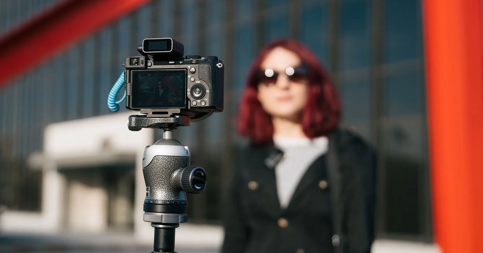 A digital camera on a tripod faces a woman with red hair and sunglasses, who stands outdoors in front of a modern glass building. The camera is in focus, while the woman and background are blurred.