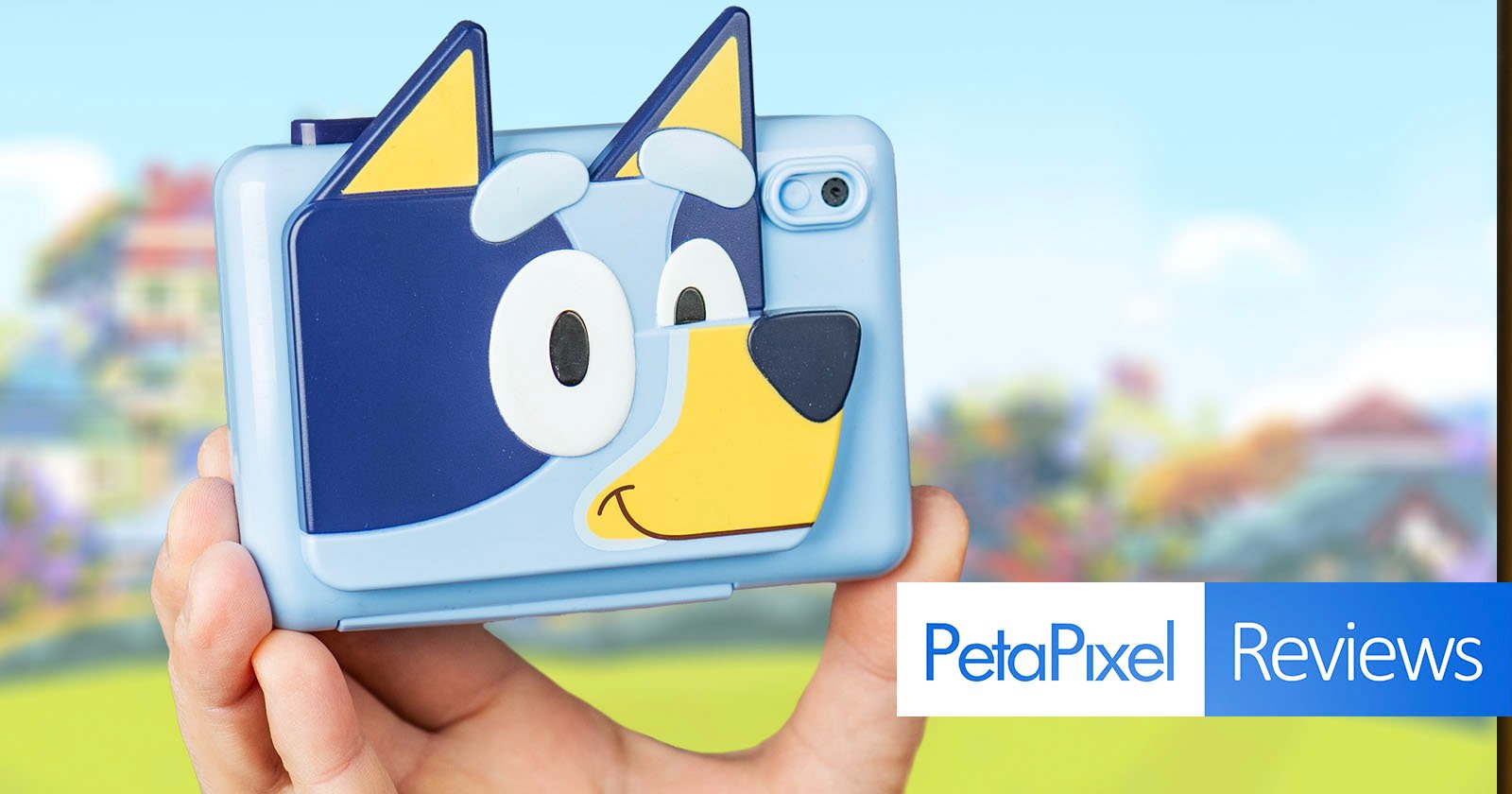 A person holds a blue camera with a cartoon dog face design on the back. The background is a blurred colorful outdoor scene. The bottom right corner shows PetaPixel Reviews text.