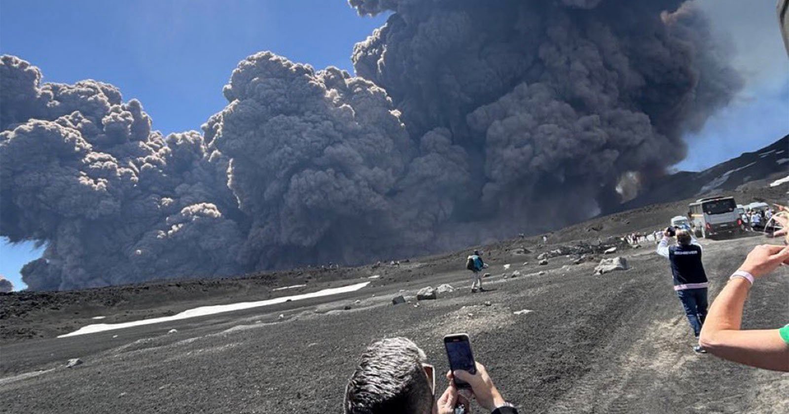 People stand on a rocky landscape taking photos as thick, dark volcanic ash clouds rise dramatically into the sky during an eruption. Cars and buses are visible on the right side.