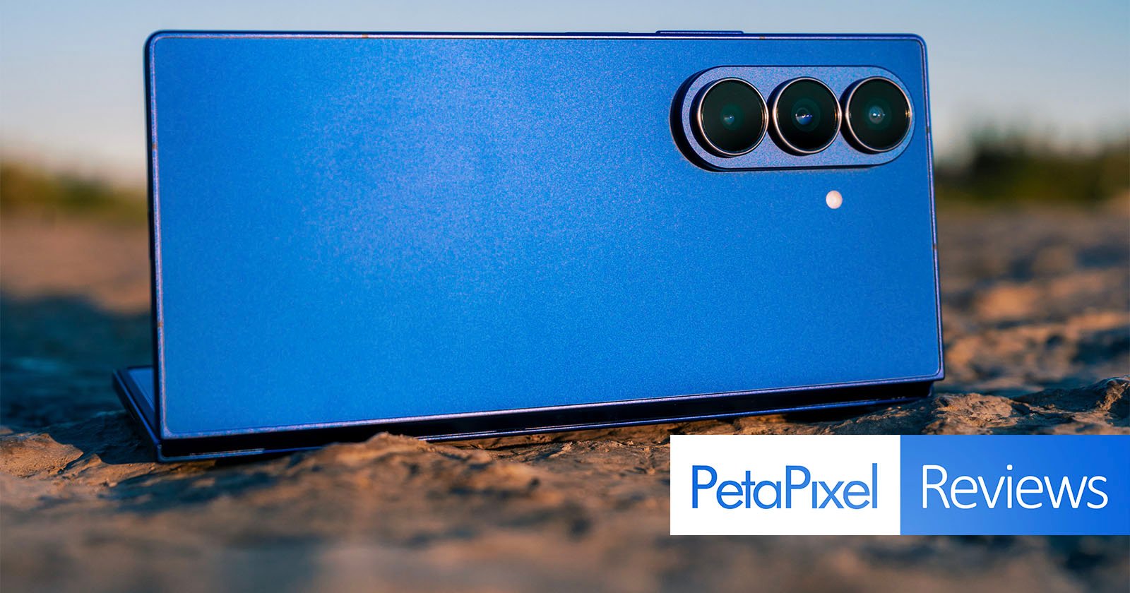 A blue smartphone with three rear cameras laying on a textured outdoor surface, with the PetaPixel Reviews logo overlaid in the bottom right corner.