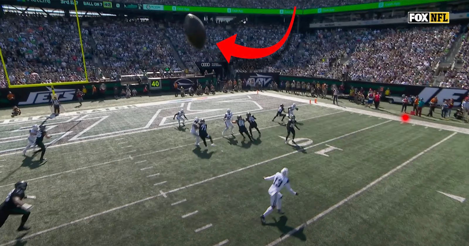 A football is in mid-air near the end zone during an NFL game, with several players looking up to catch it. The stands are full of spectators, and the Fox NFL logo appears in the top right corner.