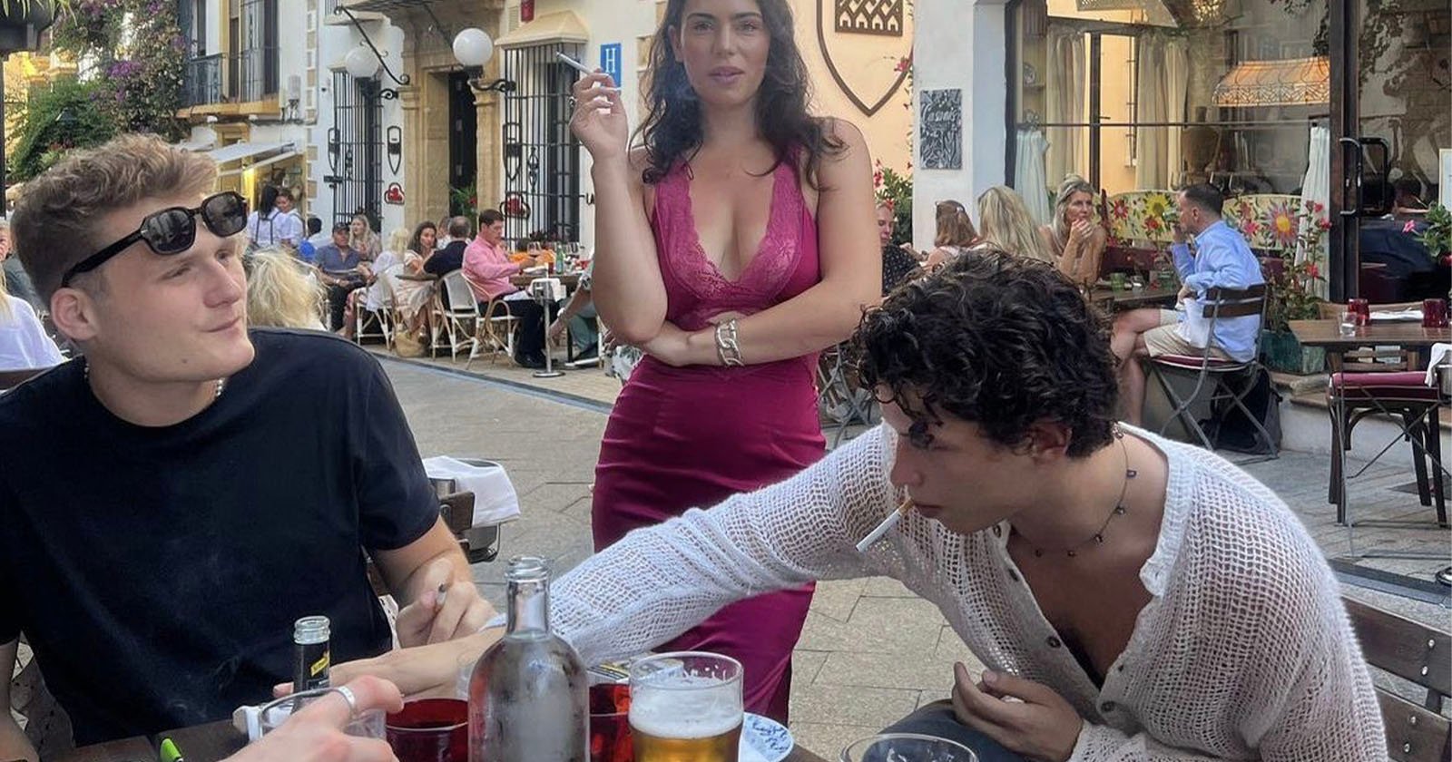 Three young adults are at an outdoor café. Two men sit at a table with drinks; one is reaching across the table, both have cigarettes. A woman in a pink dress stands behind them, also holding a cigarette, looking at the camera.
