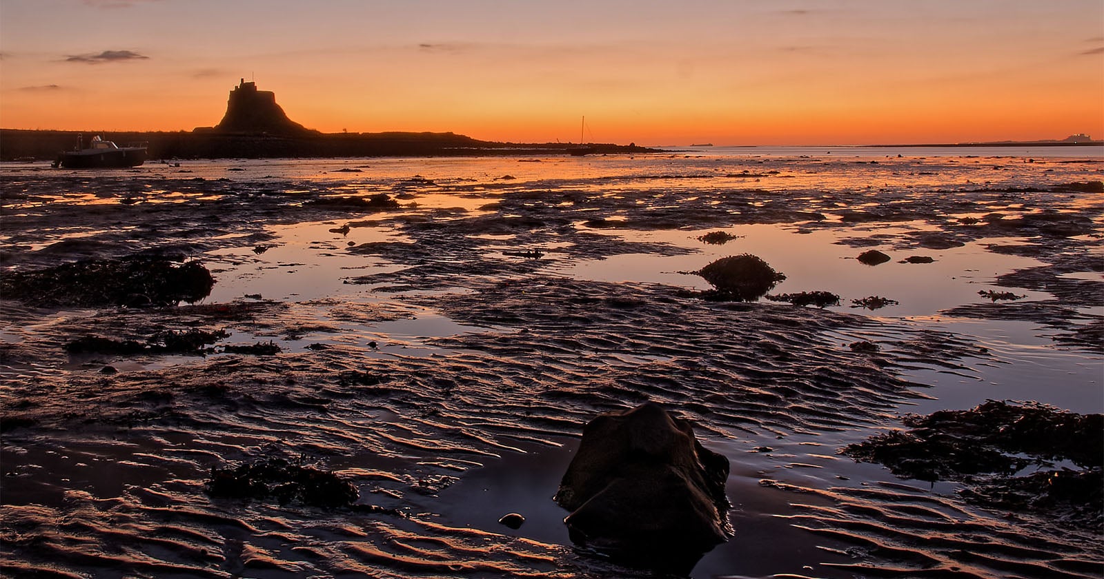 A rocky shoreline at low tide with rippled sand and puddles, silhouetted by a vibrant orange sunset. In the distance, a castle sits on a hill against the glowing sky.