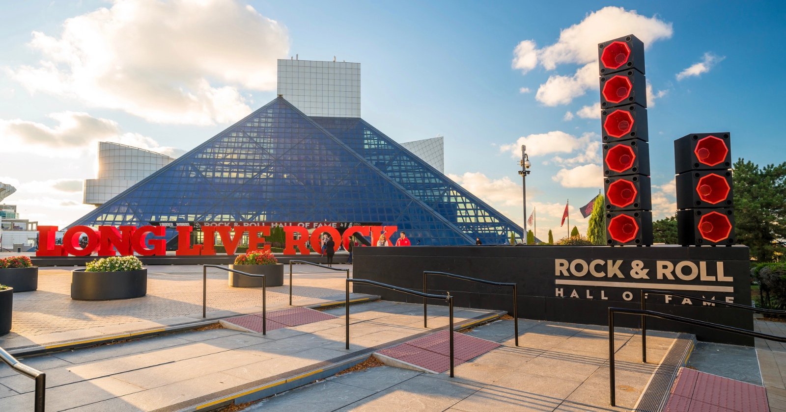 The Rock & Roll Hall of Fame in Cleveland, Ohio, features a glass pyramid structure, a large red LONG LIVE ROCK sign, and tall red speaker sculptures under a partly cloudy sky.
