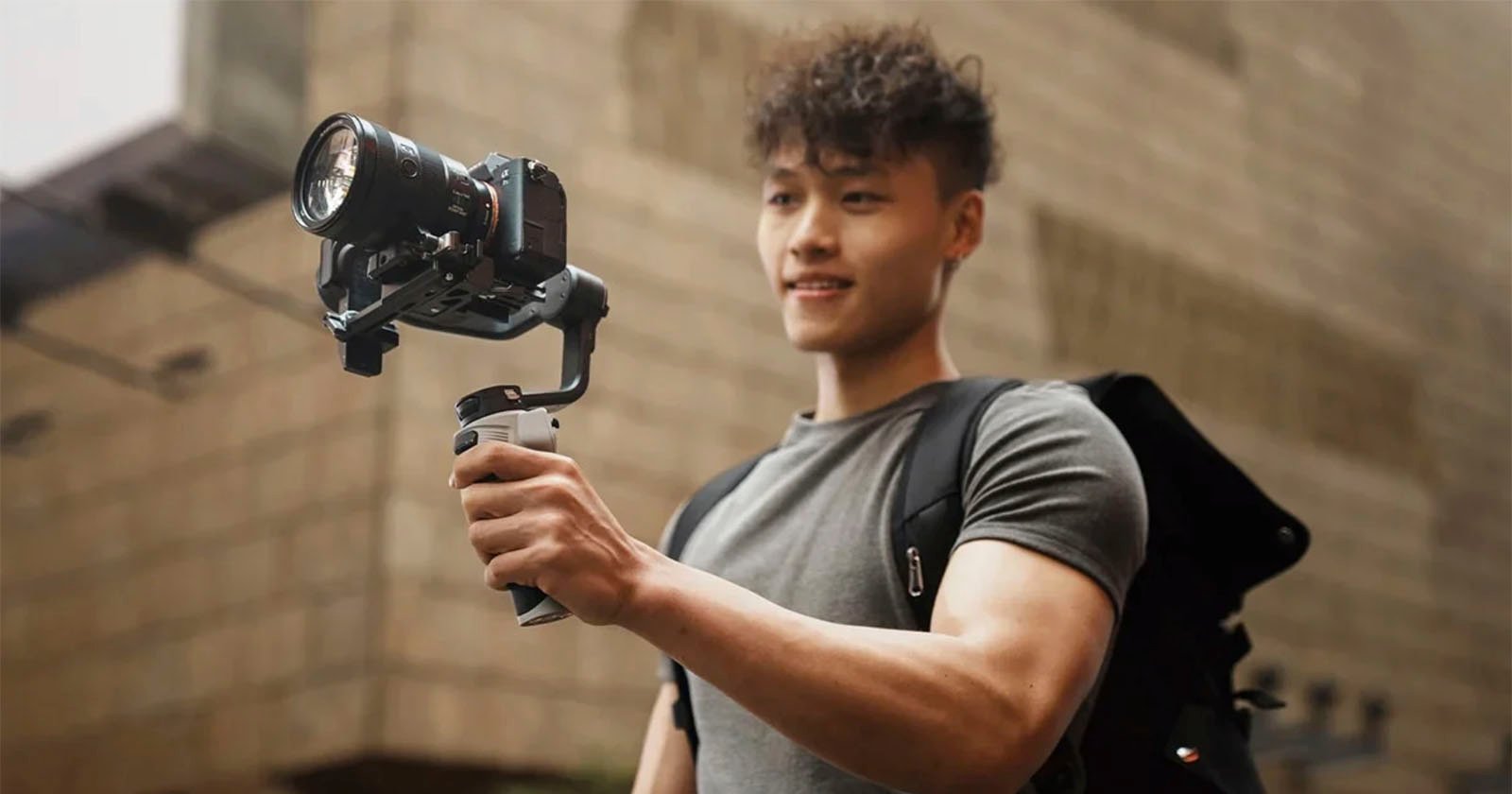 A person with short, curly hair wearing a gray t-shirt smiles while holding a professional camera mounted on a stabilizer. The person is outdoors, with a stone building in the blurred background, and has a black backpack slung over their shoulder.