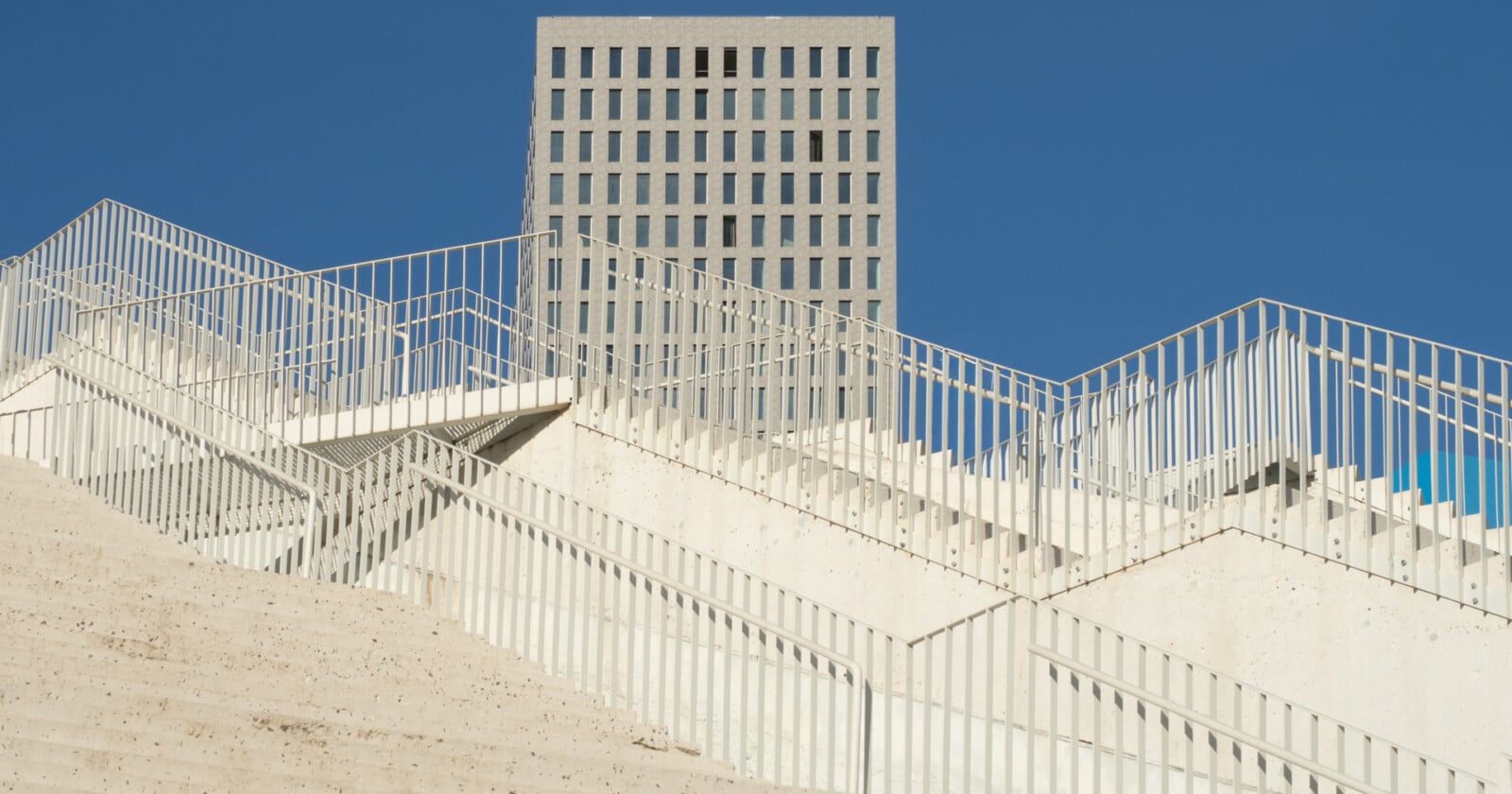 A modern, angular white staircase with metal railings rises sharply in the foreground, while a rectangular gray building with a grid of windows stands against a clear blue sky in the background.