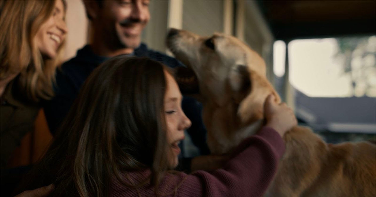 A young girl hugs a golden retriever while two adults smile in the background, all appearing happy and playful on what seems to be a porch or outdoor area.