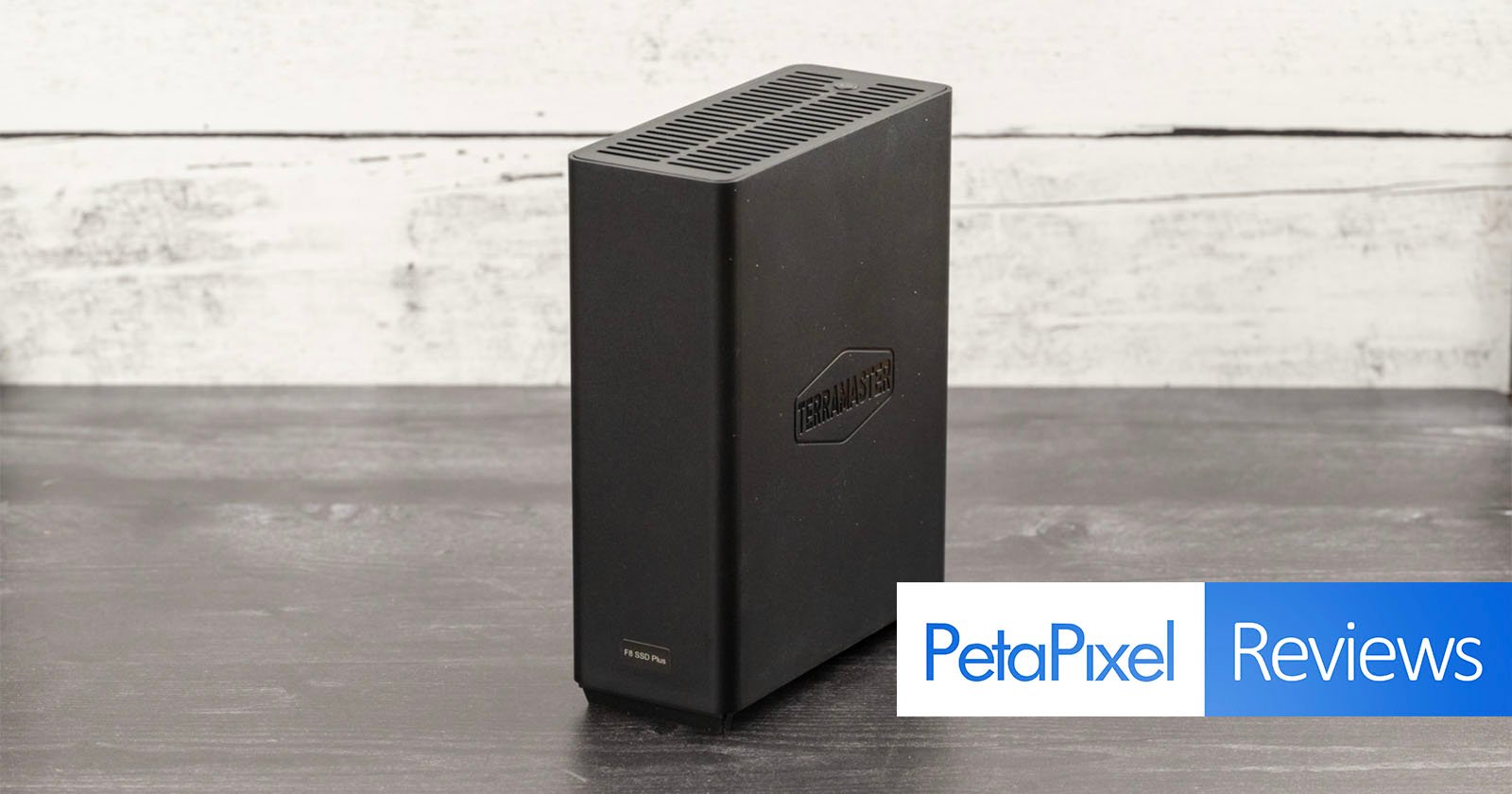 A black external hard drive sits on a wooden table against a white-washed wooden backdrop. The hard drive has the logo TerraMaster printed on it. A banner at the bottom reads PetaPixel Reviews in blue and white text.