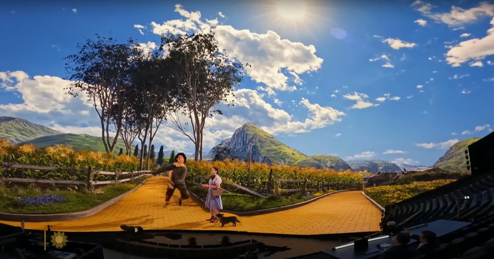 Two people in costume walk along a curved yellow brick road on a stage with a scenic digital backdrop of mountains, trees, and a bright sunny sky, evoking a scene from The Wizard of Oz.