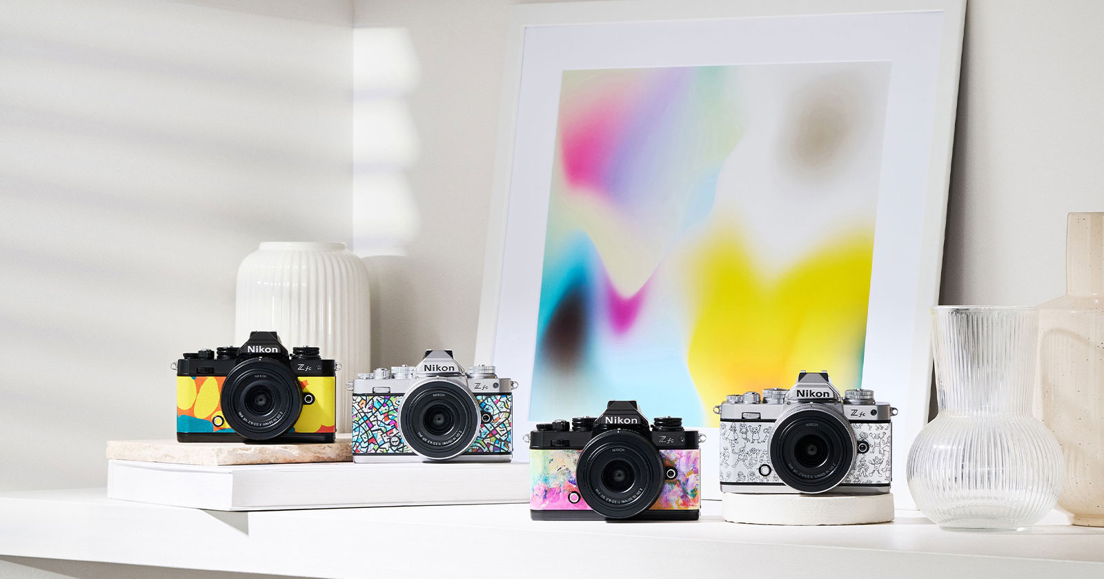 Four Nikon Z fc cameras with colorful designs are displayed on a white shelf. A vase and a glass container are on the left; to the right is another vase. A framed abstract art piece with blurred colors is on the shelf, adding a vibrant backdrop.