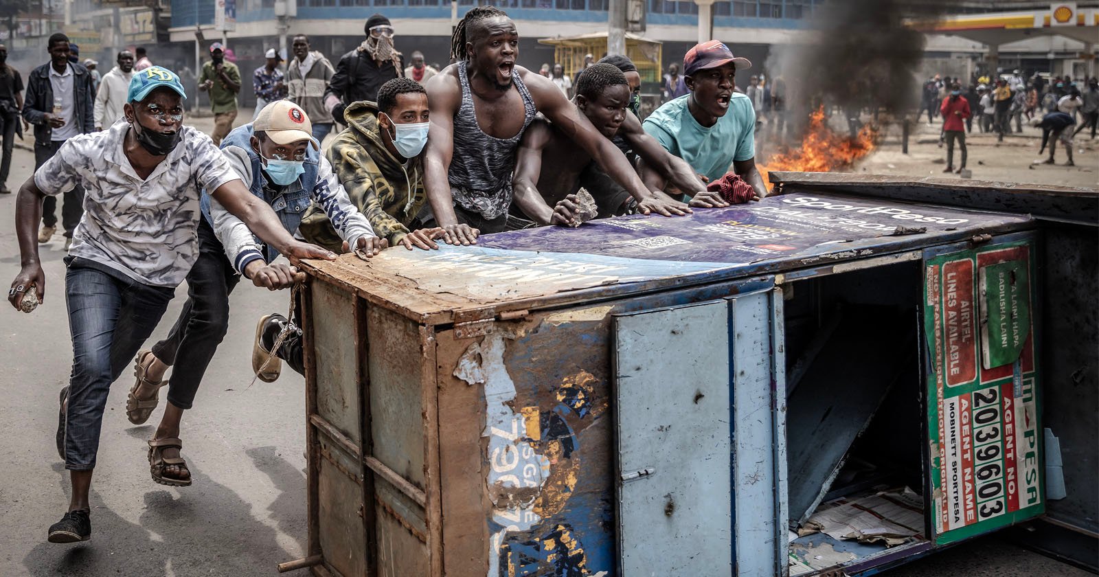 A group of men pushing an overturned metal kiosk on a street during a protest. They are wearing casual clothing and some have masks. In the background, there is smoke and a small fire, with more people gathered in the distance.