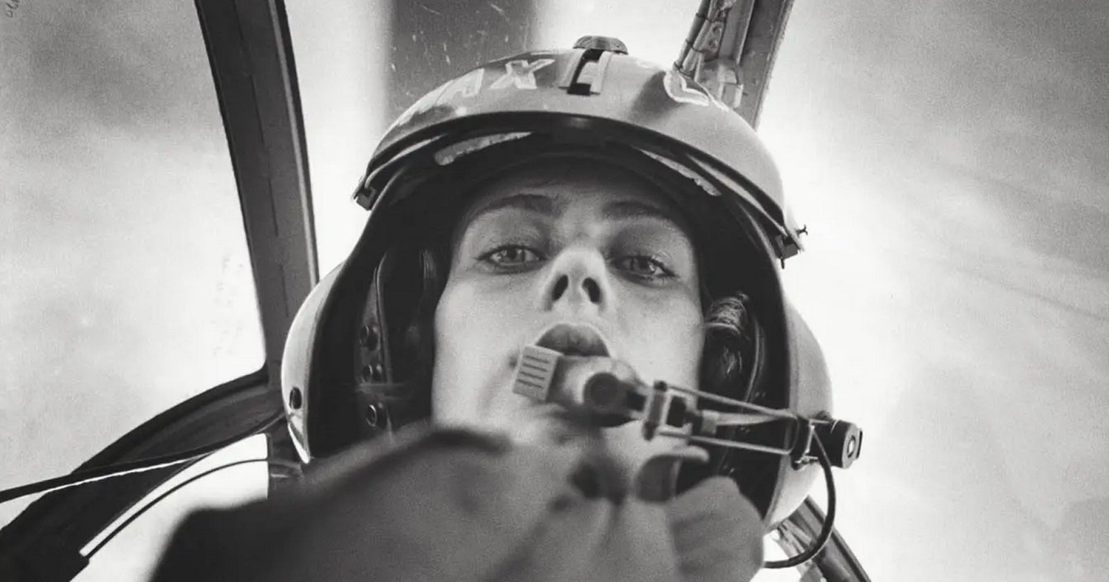 Black and white photo of a pilot wearing a helmet and headset, looking down into the camera from inside a cockpit, with blurred background and cockpit structures visible.