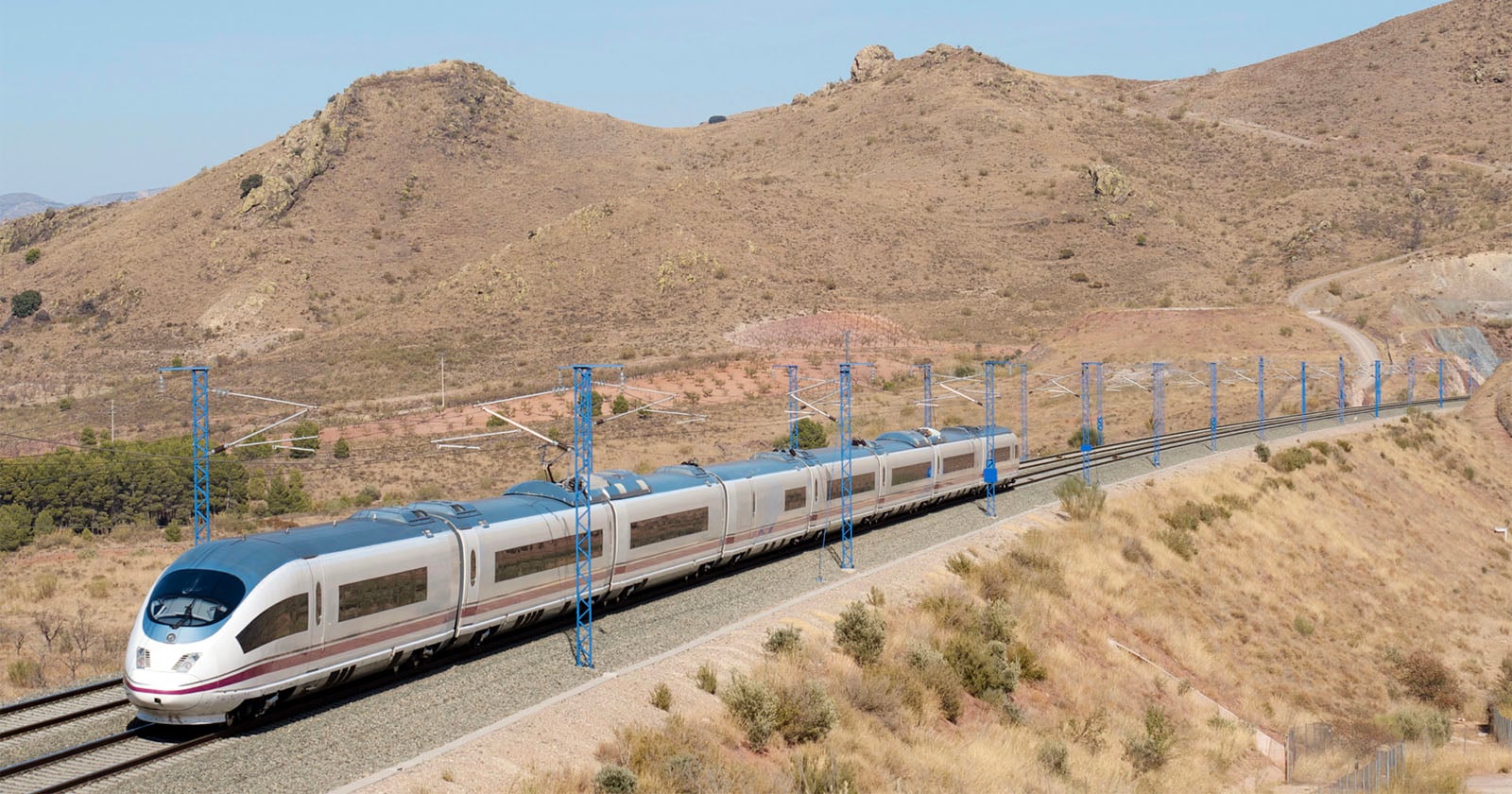 A modern high-speed train travels through a dry, hilly landscape with brown grass and scattered shrubs under a clear sky. Blue electric poles line the railway track.