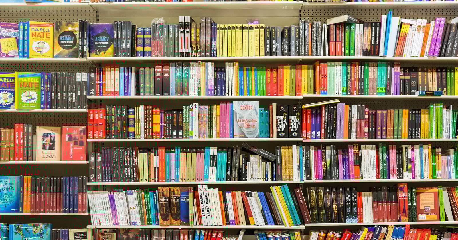 A bookstore shelf filled with colorful, neatly arranged books of various sizes and genres, including novels, graphic novels, and children's books, organized mainly by color and height.