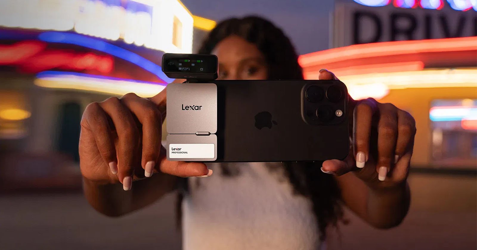 A person holding a smartphone with an attached Lexar external memory module and an additional device over the camera, possibly for enhanced photography or video. The background is blurred with neon lights, suggesting an urban night scene.