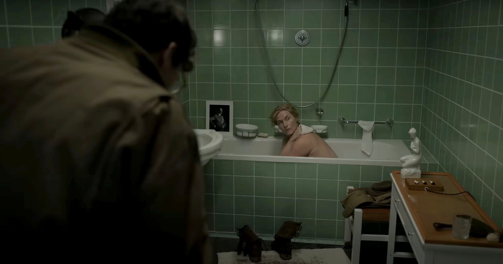 A person is in a bathtub filled with water in a green-tiled bathroom, turning to look at someone standing nearby. Various items such as towels, clothes, and grooming tools are scattered around, including a pair of boots on a mat in front of the tub.