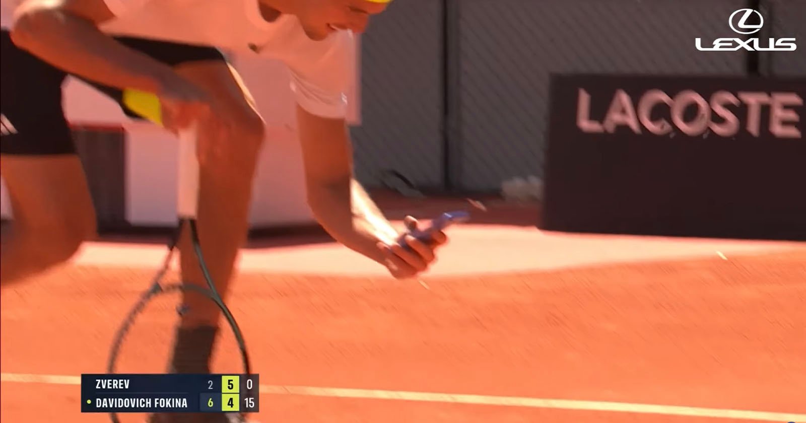 A tennis player in a white shirt and yellow headband bends down on a clay court, holding a phone and racket. Score graphics and the Lexus logo appear on the image.