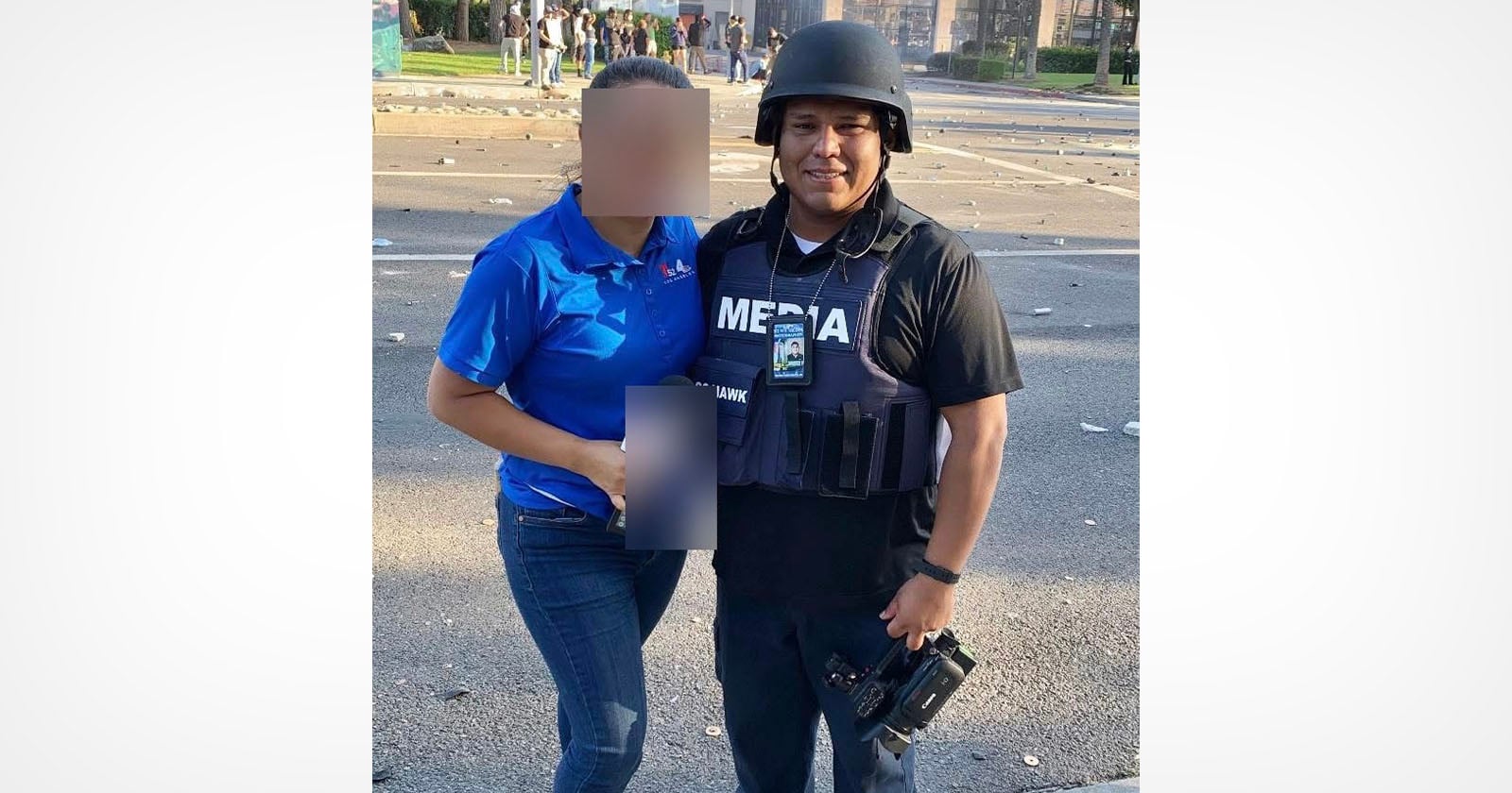 A woman in a blue shirt stands next to a man wearing a helmet, a MEDIA vest, and holding a camera. Their faces are blurred. Debris is scattered on the street in the background.