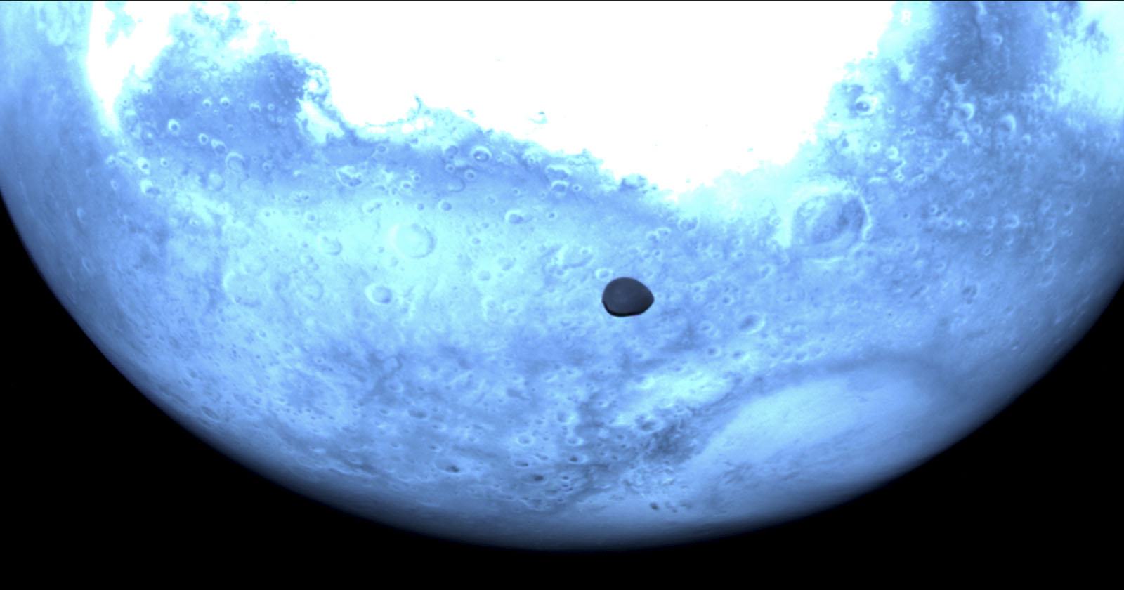 A close-up view of a blue-tinted planet surface with craters and rough terrain. A small, dark circular shadow is visible on the planet's surface, indicating an eclipse or transit event.