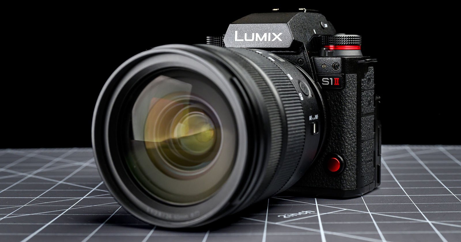 A close-up of a Panasonic Lumix S1 II digital camera with a large lens, placed on a grid-patterned surface against a black background.