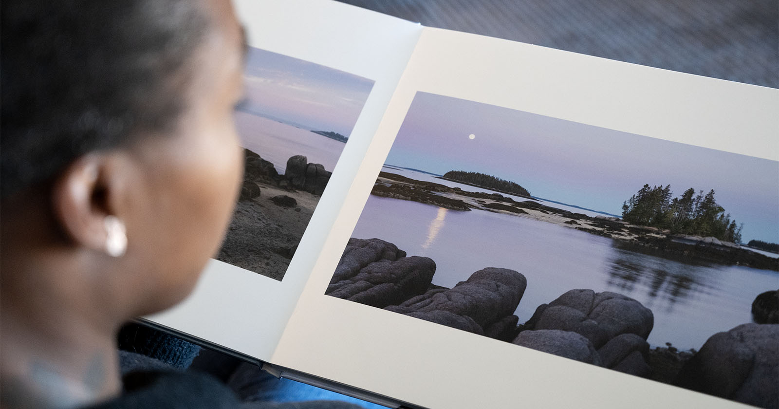 A person is looking at an open photo book displaying serene landscape images of a rocky shoreline and calm waters under a pastel sky. A small island with trees is visible in the distance.