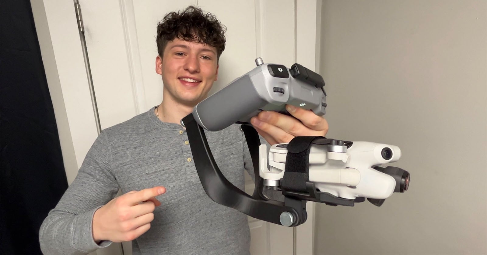 A smiling young man in a gray shirt holds a folded white drone attached to a black handheld stabilizer and points at it, standing indoors against a white door and light-colored wall.