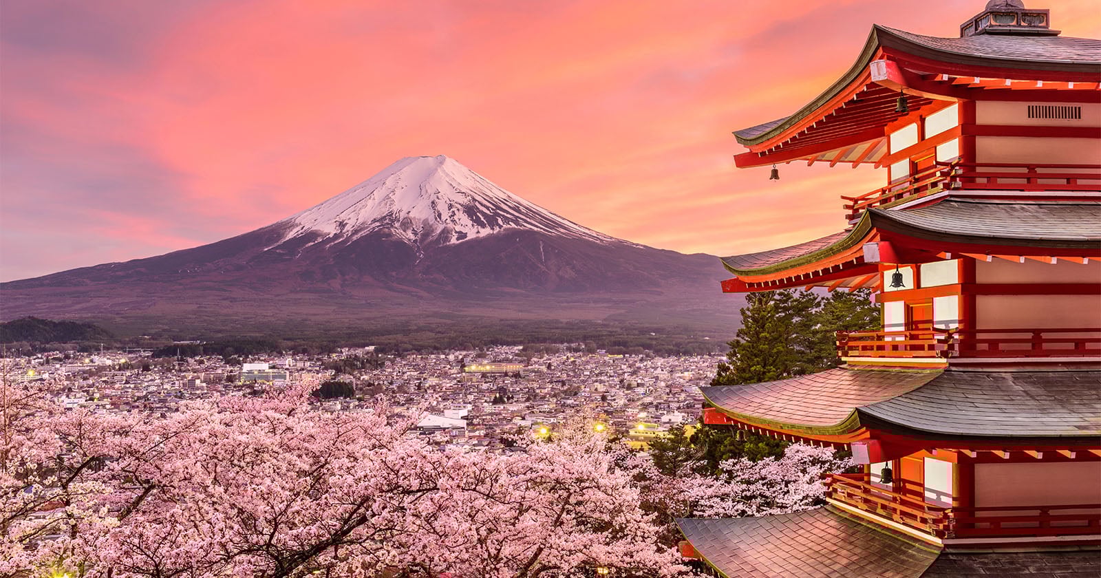 A traditional Japanese pagoda stands among cherry blossoms, with Mount Fuji and a town in the background under a vibrant pink and orange sunset sky.