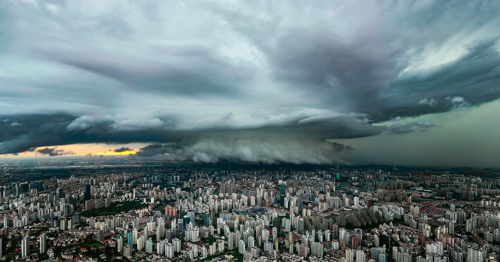 A dramatic, dense storm cloud hovers over a sprawling city skyline at sunset, with tall buildings below and dark, turbulent clouds dominating the sky.