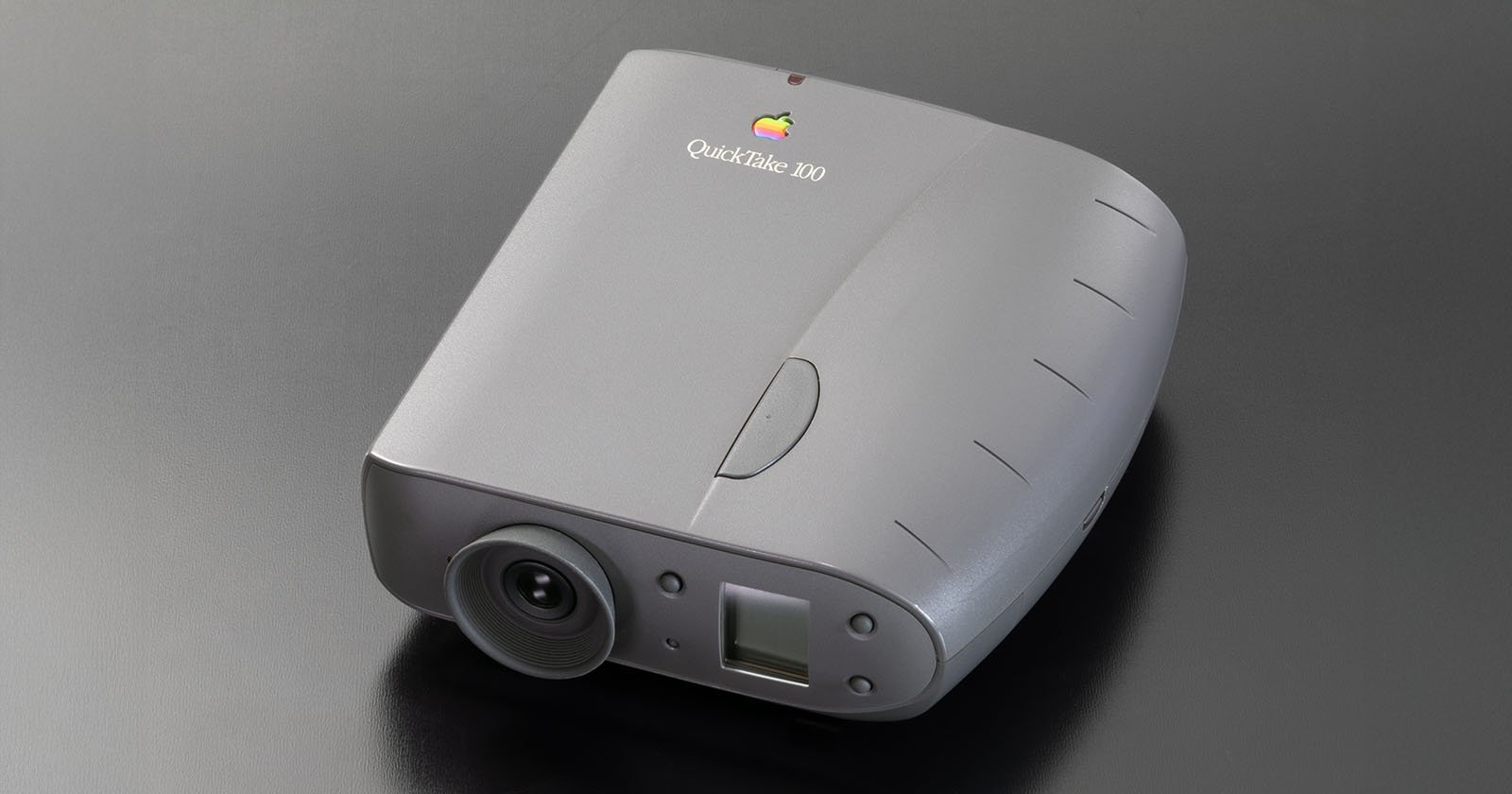 A gray Apple QuickTake 100 digital camera with a rounded body, lens on the front, and Apple logo on top, placed on a sleek dark surface.