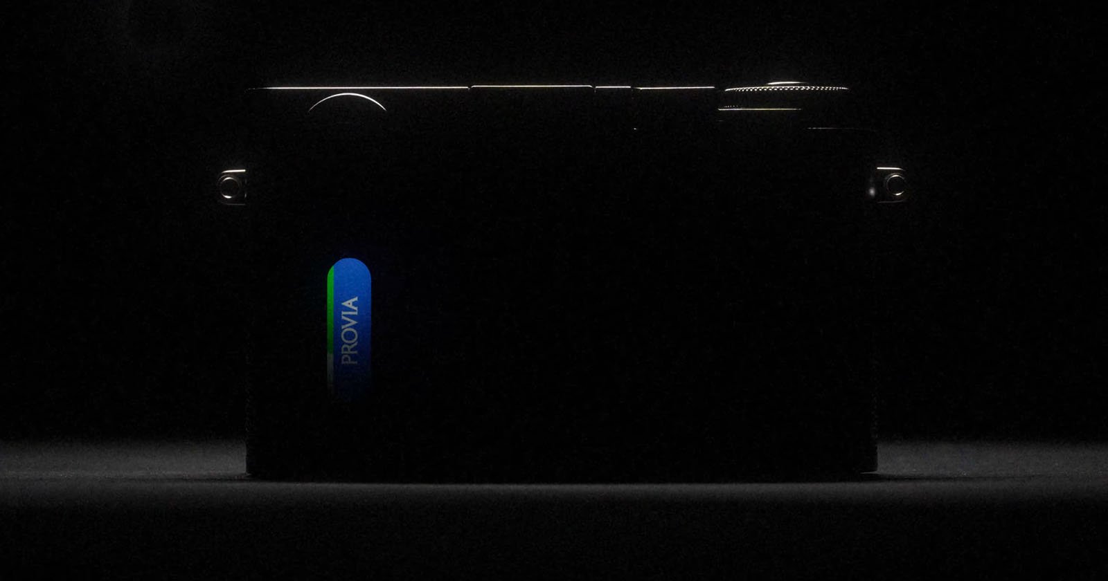 A dark, low-light image of a camera, mostly in shadow, with a bright blue and green label reading PROVIA illuminated on its side. The camera's outline and dials are just barely visible.