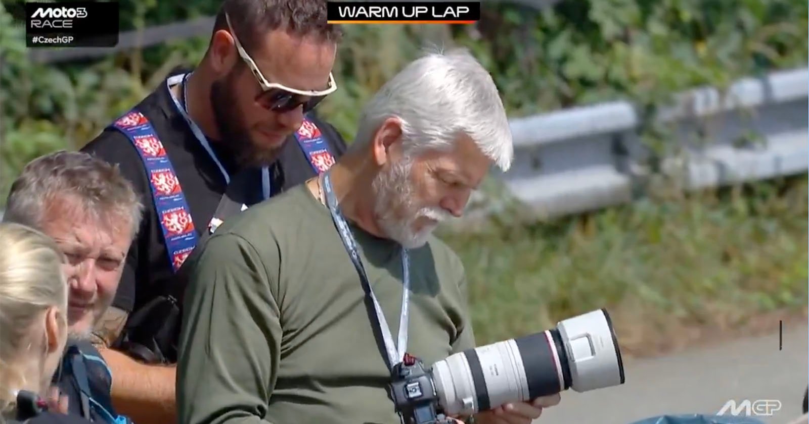 Two men with cameras, one with a large telephoto lens, review photos outdoors near a racetrack during a MotoGP event. A WARM UP LAP graphic is displayed at the top of the image.