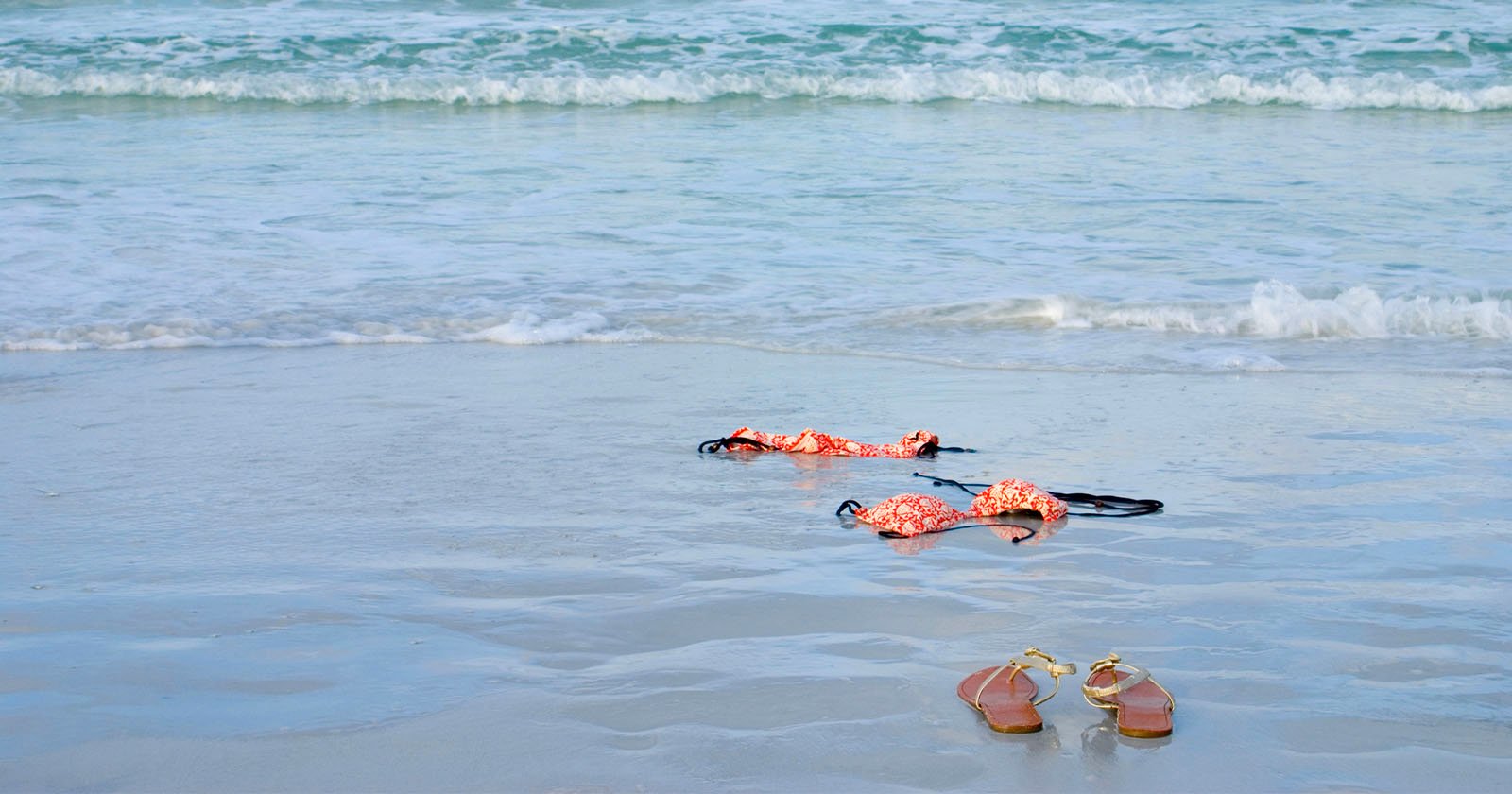 A pair of sandals and a floral bikini lie on the wet sand near the shoreline, with gentle ocean waves approaching in the background.