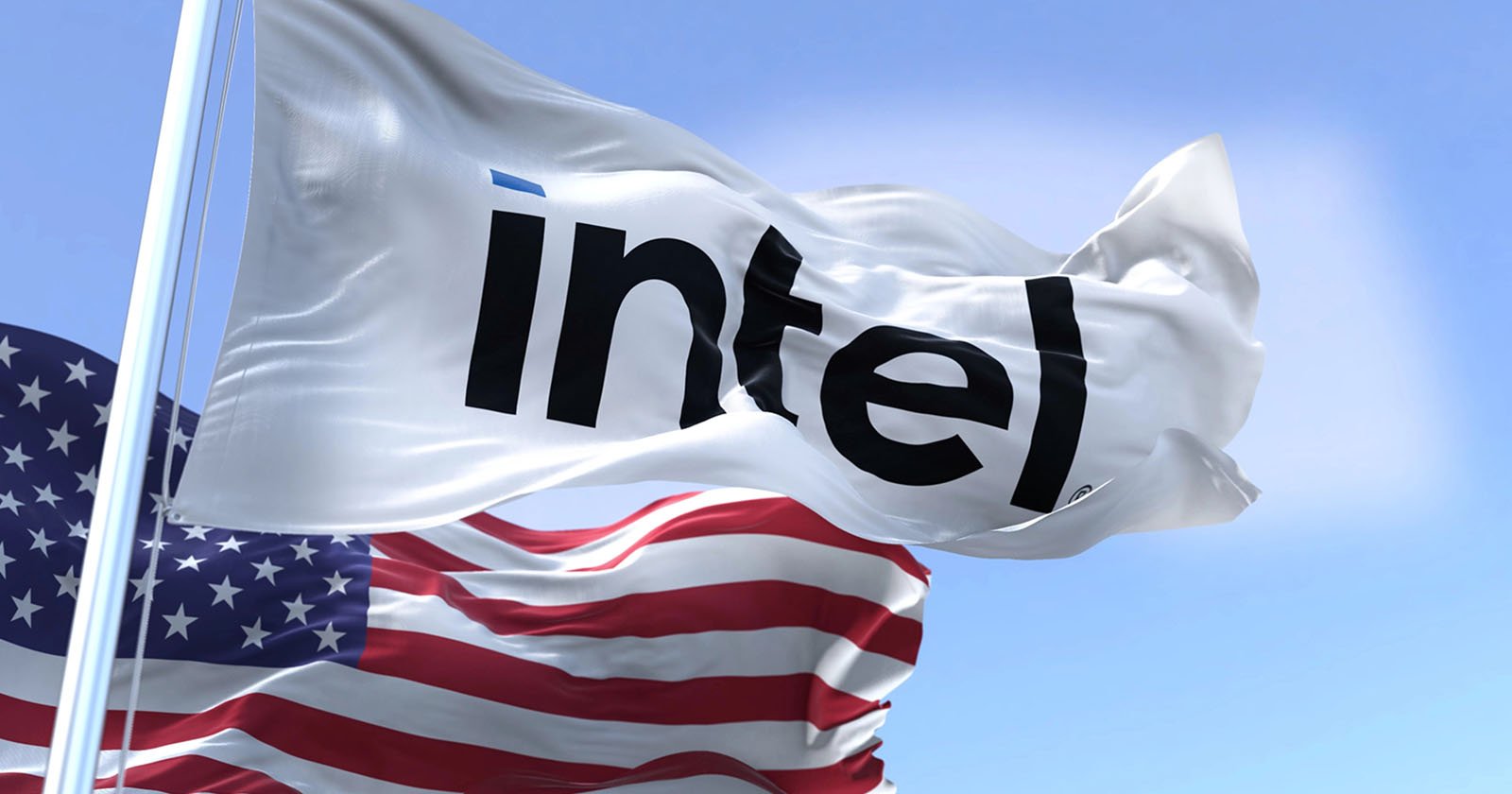 A white flag with the black Intel logo waves in the wind next to an American flag against a blue sky with clouds.