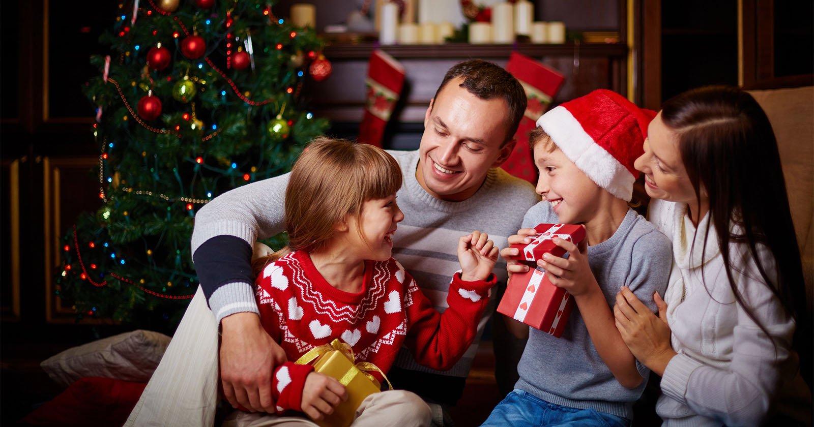 A family of four sits together by a decorated Christmas tree. The two children, a boy wearing a Santa hat and a girl in a festive sweater, exchange wrapped gifts. The parents smile warmly, enjoying the holiday atmosphere.