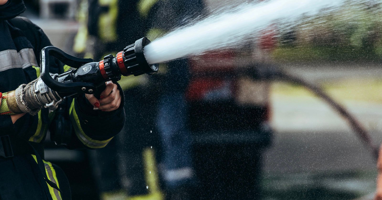 A firefighter wearing protective gear sprays water from a hose, with water forcefully streaming out. The background is blurred, showing other firefighters and equipment.