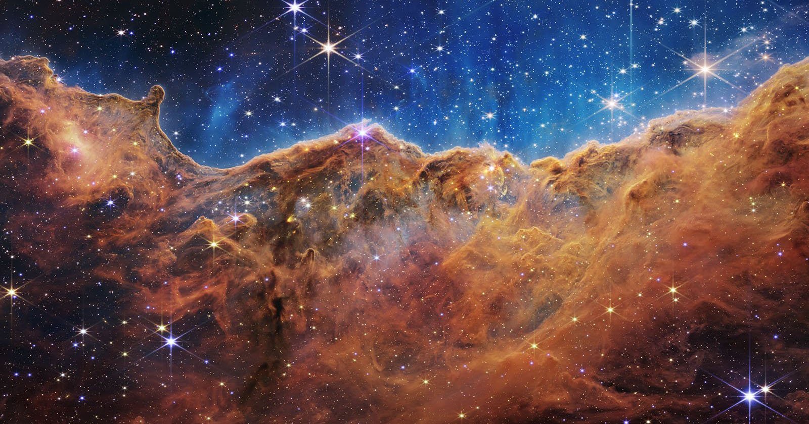 A colorful image of a star-forming region in space shows bright stars scattered across a blue and black sky above glowing, orange-brown clouds of gas and dust, resembling a mountainous landscape.