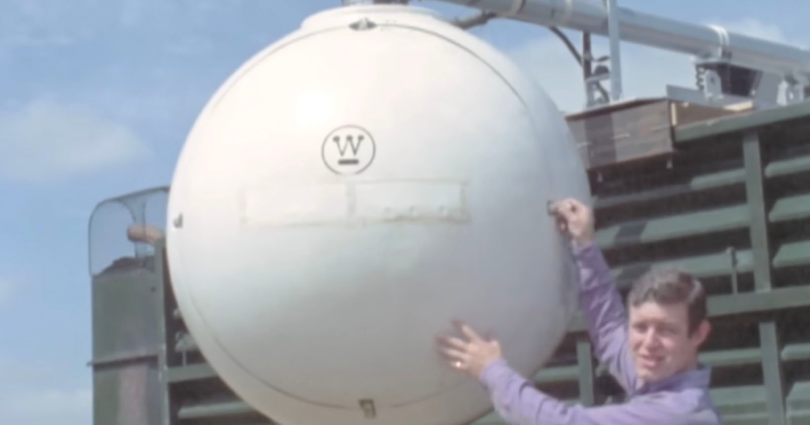 A man in a purple shirt stands next to a large, spherical white tank with a W logo on it, mounted on industrial equipment outdoors under a blue sky.