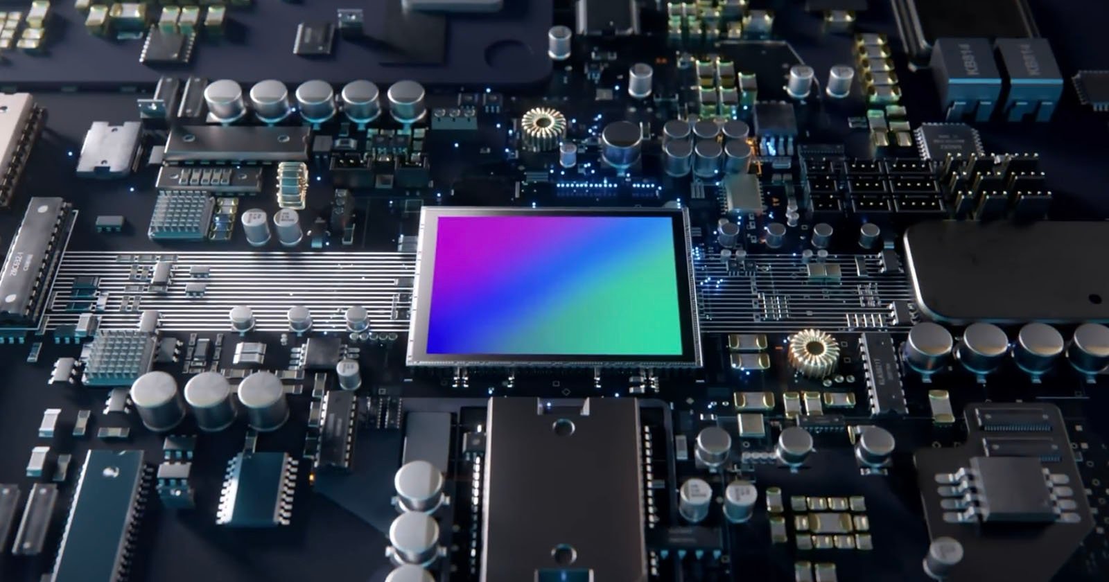A close-up view of an electronic circuit board featuring various components and a central square chip with a colorful gradient surface, surrounded by capacitors, resistors, and intricate wiring.