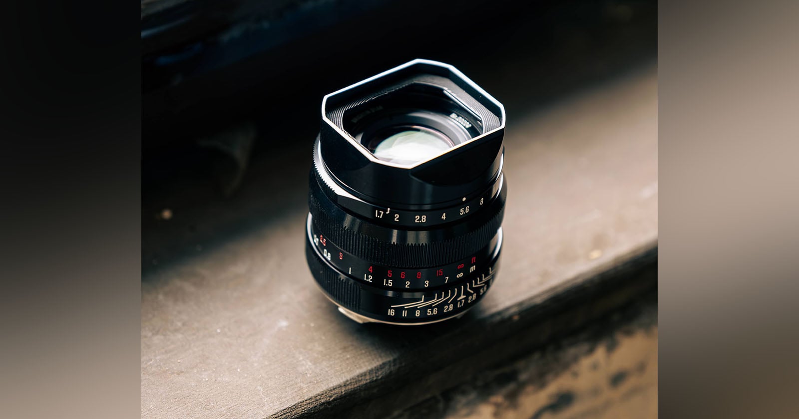 A close-up shot of a black camera lens with white and red markings, placed on a sunlit wooden surface. The lens hood is attached, and the background is softly blurred.