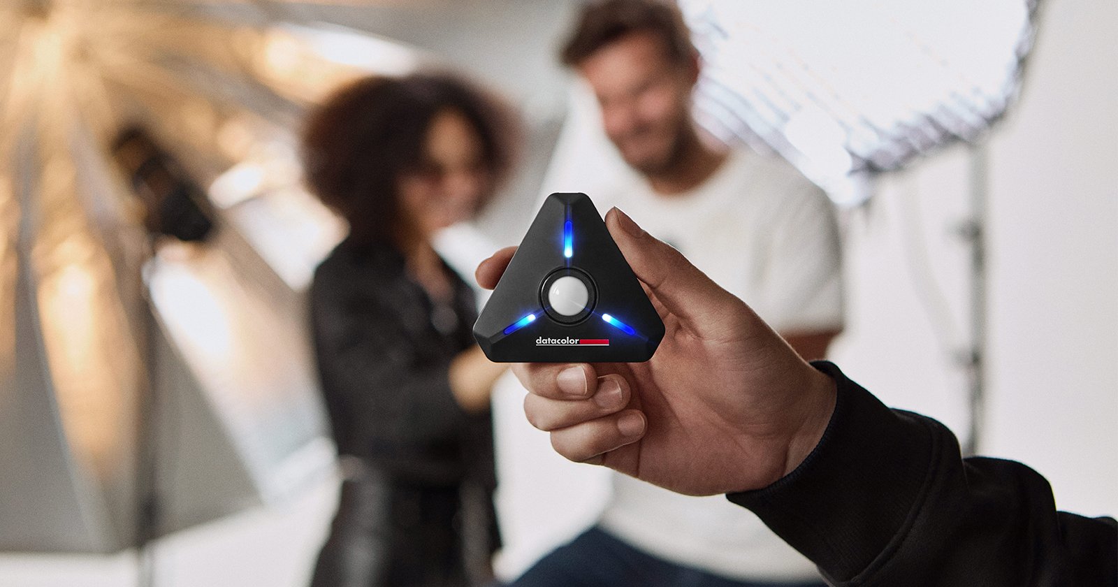 A person holds a black, triangular device with blue lights, focusing on it. Behind, two people are blurred, with professional lighting equipment in the backdrop. The device is central to the image.