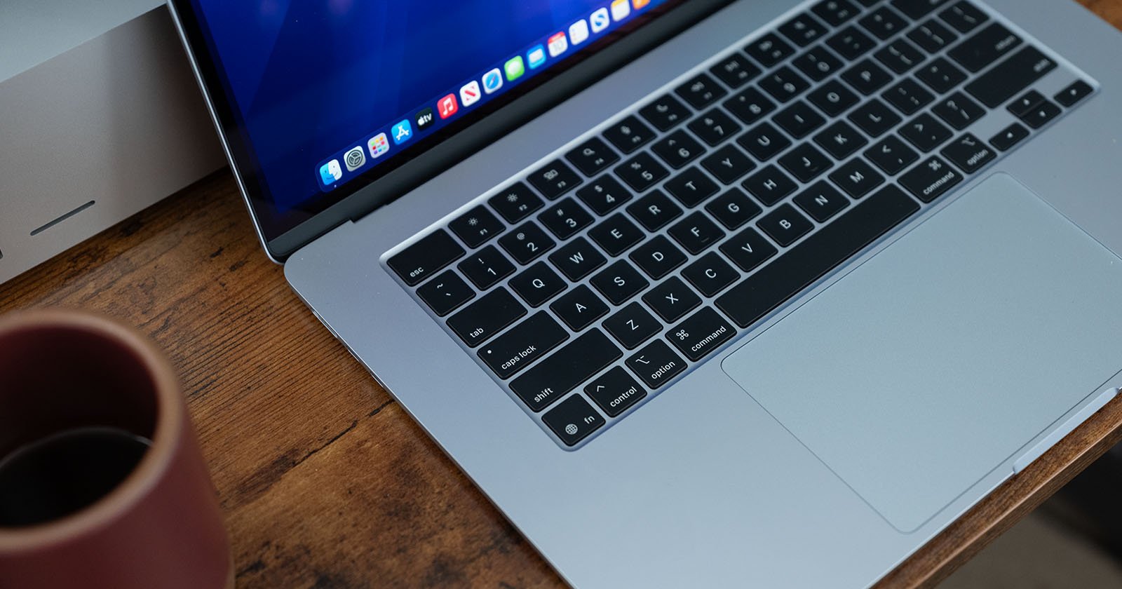A close-up of a laptop keyboard and touchpad on a wooden desk, with a brown mug of coffee and another electronic device visible in the background.