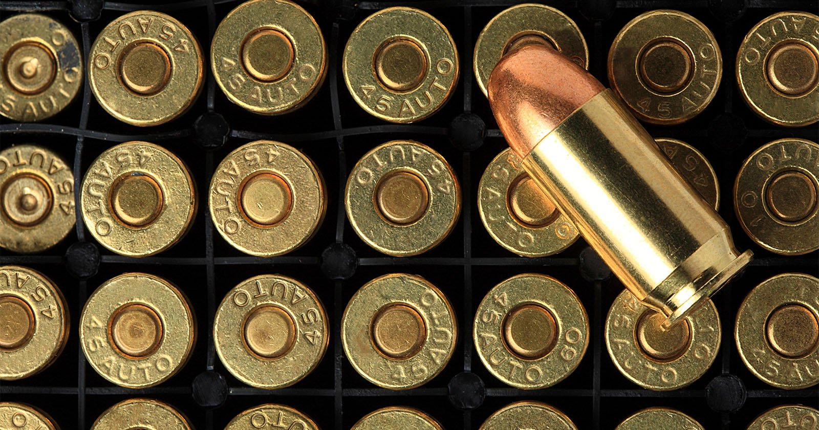 Close-up of a tray filled with .45 Auto pistol cartridges, showing the brass casing with 45 AUTO markings. One cartridge is positioned horizontally above the others, displaying its full length and copper jacketed bullet. The background is black.