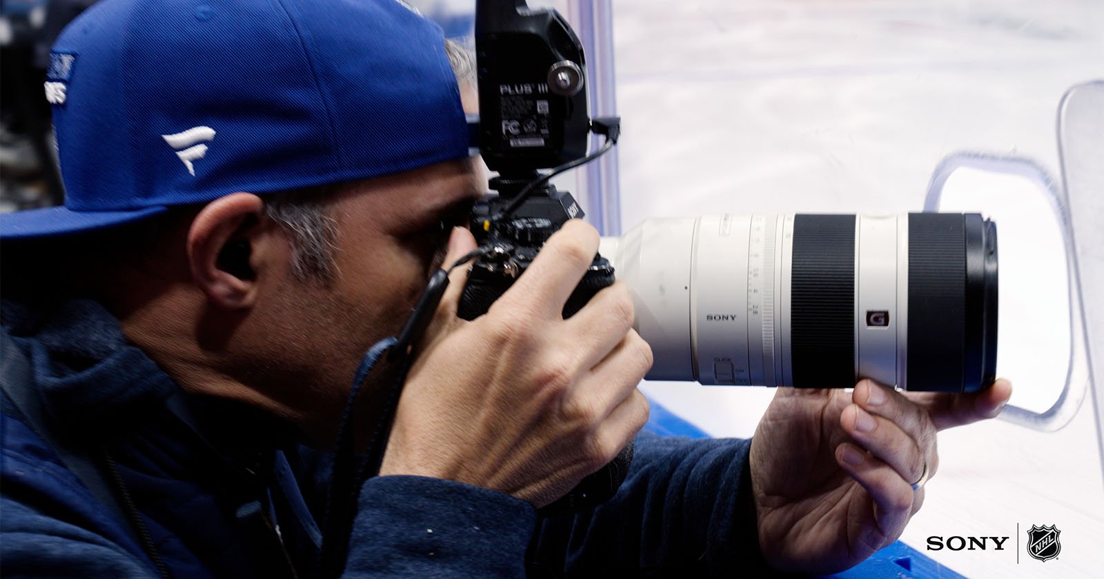 A man wearing a blue cap and jacket holding a Sony camera with a large telephoto lens, aiming to take a photo. Sony and NHL logos are visible in the bottom right corner.
