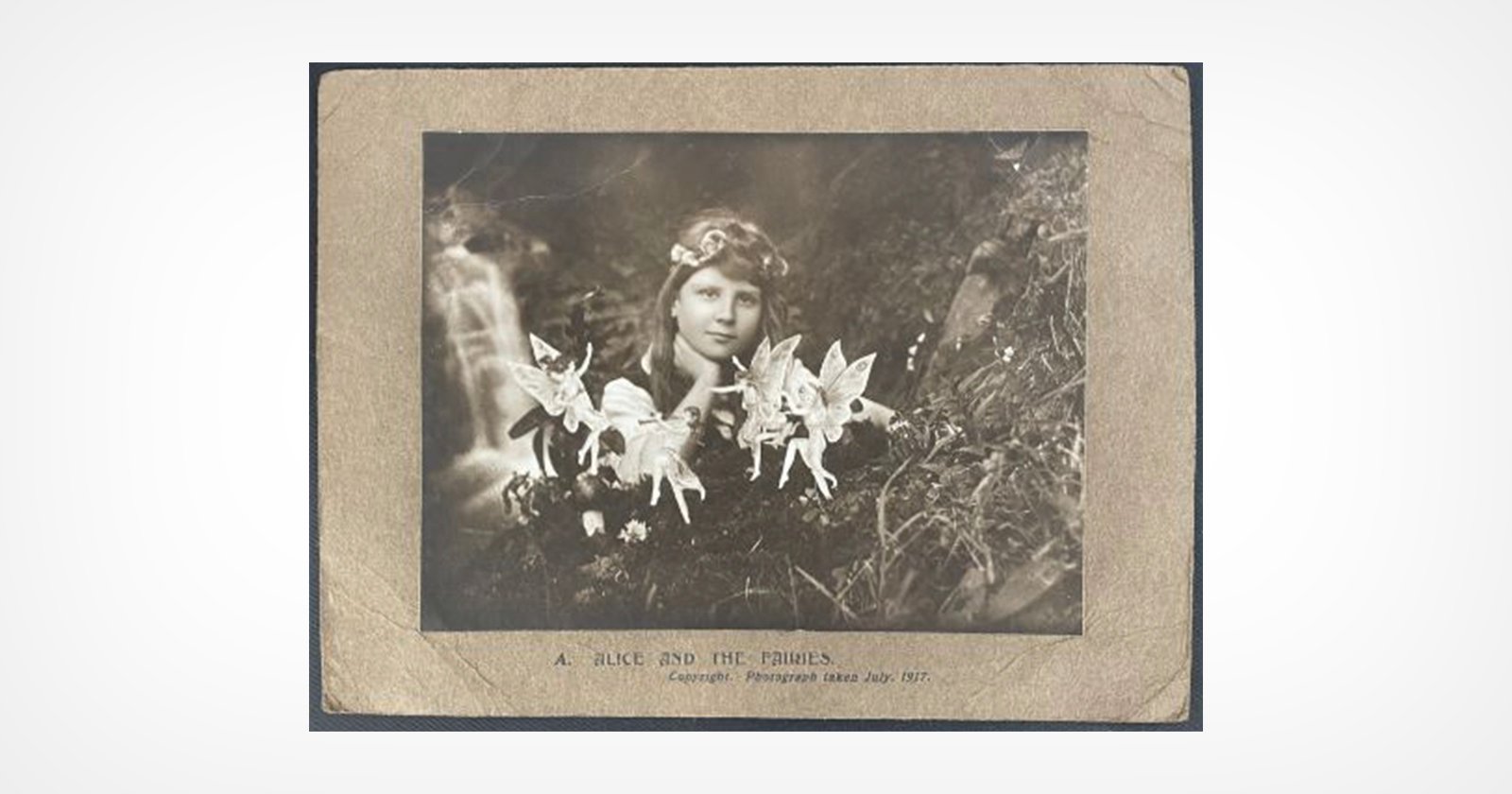 A vintage black-and-white photo of a girl outdoors with paper cutout fairies placed in front of her, blending into the natural background. The caption reads, A Slide and the Fairies.