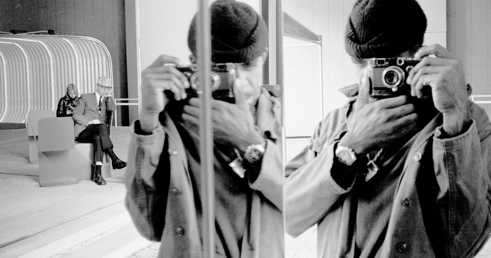 A person in a coat and beanie takes a photo with a camera in front of a mirror, creating a reflection. In the background, an older couple sits on a bench near a modern, curved structure. The image is black and white.