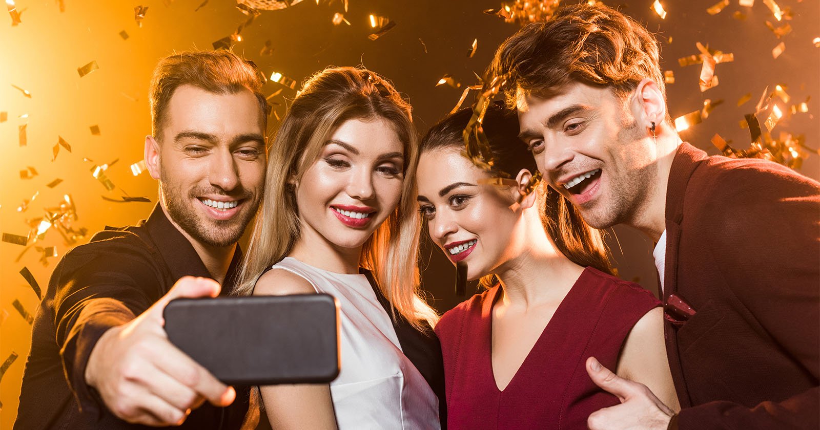 Four people are taking a selfie together at a celebration, smiling and appearing joyful. The background is filled with golden confetti and warm lighting, creating a festive atmosphere.