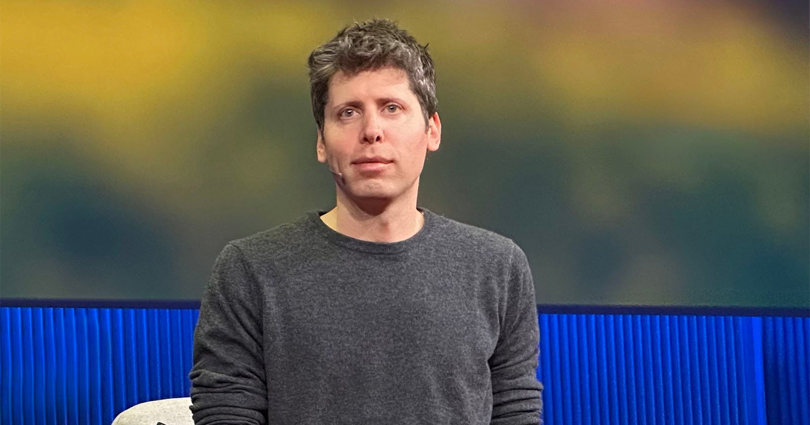 A man with short, tousled brown hair wearing a gray sweater sits in front of a blurred background with blue and yellow tones.