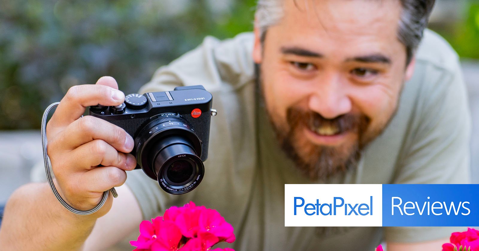 A man with a beard is smiling as he takes a close-up photo of pink flowers with a black camera. The background is blurred, highlighting the flowers and the photographer. There is an overlay with the text PetaPixel Reviews at the bottom right corner.